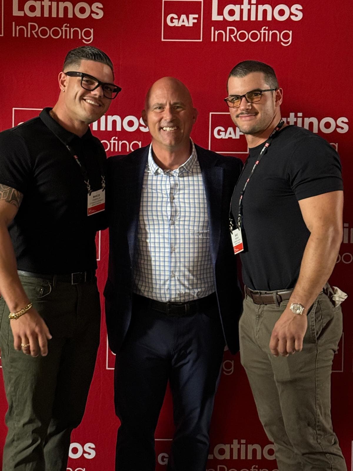 Tres hombres posan juntos frente a un fondo rojo de GAF Latinos InRoofing.